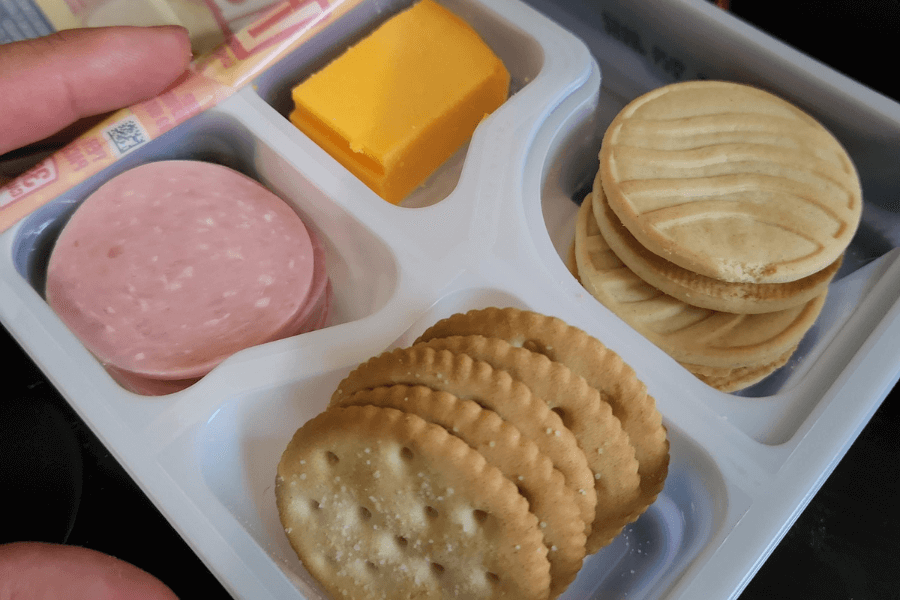 A divided plastic lunch tray containing a stack of round crackers, a few slices of deli-style meat (likely bologna), a thick slice of cheddar cheese, and two sandwich cookies with a swirl design on top.