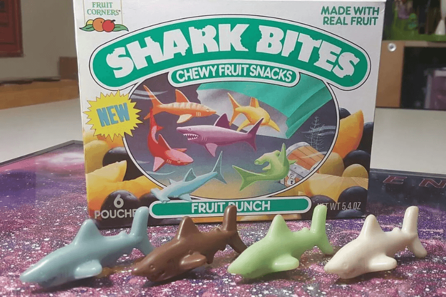 A box of Shark Bites fruit snacks in the fruit punch flavor sits behind five toy shark figures resembling the snack shapes. The retro packaging shows colorful illustrated sharks and tropical fruit, promoting that it’s made with real fruit and contains six pouches.