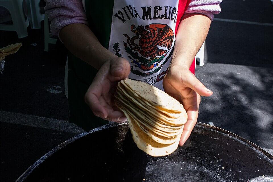 A person wearing a "Viva Mexico" apron holds a freshly made stack of warm corn tortillas over a hot griddle. The tortillas are golden brown with slightly charred edges, and sunlight casts a glow on their texture.