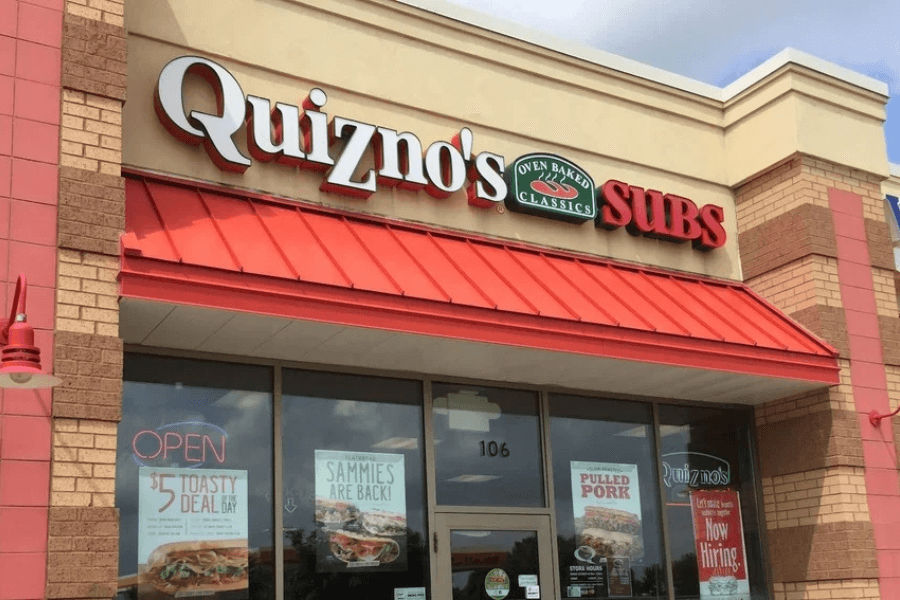 The image showcases a Quizno's restaurant with its recognizable red and tan exterior, featuring signs promoting toasted subs and specials. Its bright, inviting storefront reflects the chain’s focus on unique, oven-toasted sandwiches.