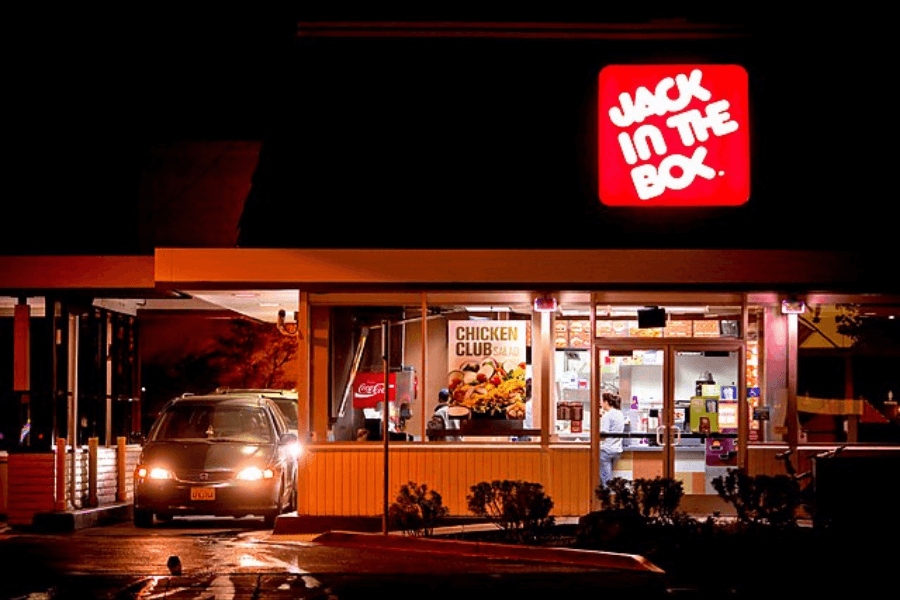 The image shows a warmly lit Jack in the Box restaurant at night, featuring a glowing red and white sign and a drive-thru with a car in line, evoking memories of late-night cravings.