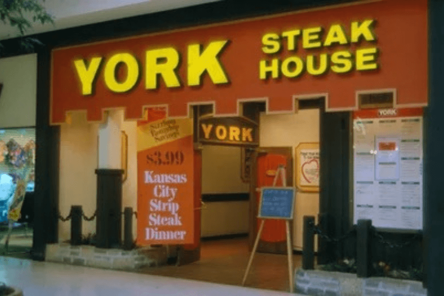 This image shows the entrance of York Steak House, featuring bright yellow signage, a warm brick-red exterior, and a vintage menu board advertising a Kansas City Strip Steak Dinner for $3.99, evoking classic steakhouse vibes.