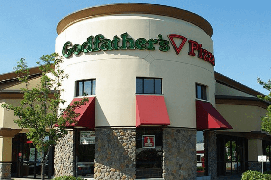 This image shows the exterior of a Godfather's Pizza restaurant, characterized by its tan stucco walls, stone accents, bold green-and-red logo, and red awnings, creating a welcoming and classic Italian eatery vibe.