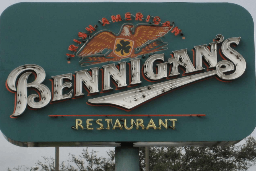 The image shows a Bennigan's restaurant sign featuring the words "Irish American" with an eagle holding a clover emblem above the iconic Bennigan's logo, evoking nostalgia for the classic Irish-American dining experience.