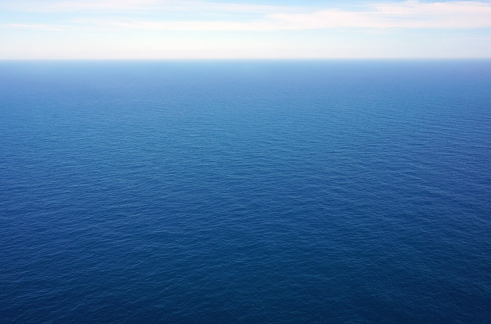 Endless expanse of calm, deep blue ocean stretching out to a pale blue horizon under a lightly clouded sky. The water surface is smooth with gentle ripples, creating a serene and tranquil scene.