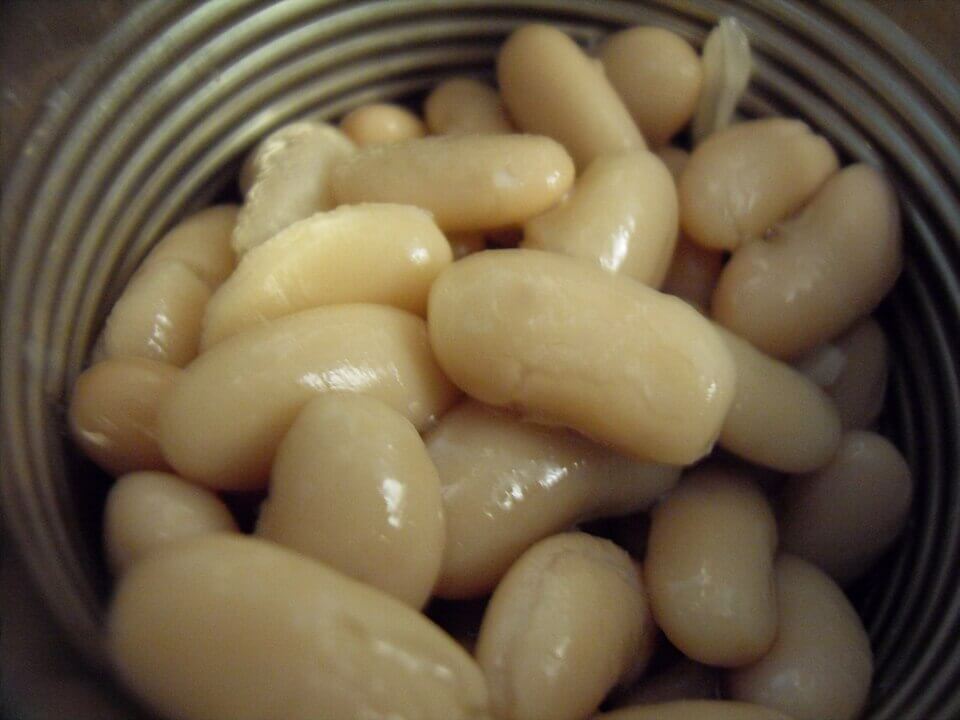 Close-up view of canned white beans packed tightly inside a metal can, with a shiny, slightly slimy texture and light beige color reflecting the surrounding light. The ridged interior of the can is partially visible around the beans.