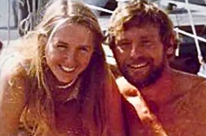 Smiling man and woman sitting closely together on a boat, both sun-kissed and relaxed, with the woman wearing a shell necklace and the man shirtless with tousled hair and a beard. Sailboat rigging and deck are visible in the background, suggesting a warm, seaside setting.