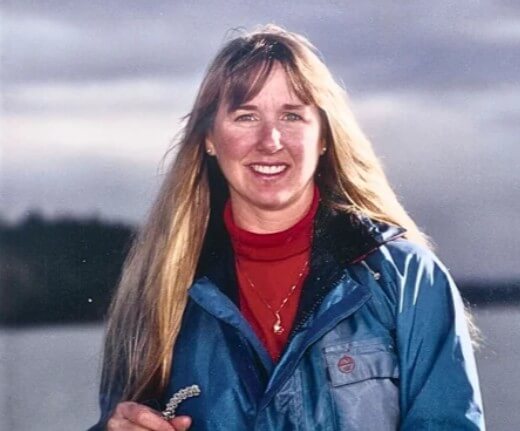 Smiling woman with long blonde hair standing outdoors by a body of water, wearing a blue jacket over a red turtleneck and holding a rope in her hand. The background features a cloudy sky and dark, forested shoreline.
