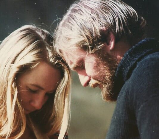 Close-up of a blonde woman and a bearded man leaning their foreheads together, eyes cast downward in a quiet, emotional moment. The lighting is soft, creating an intimate and tender atmosphere between the two.