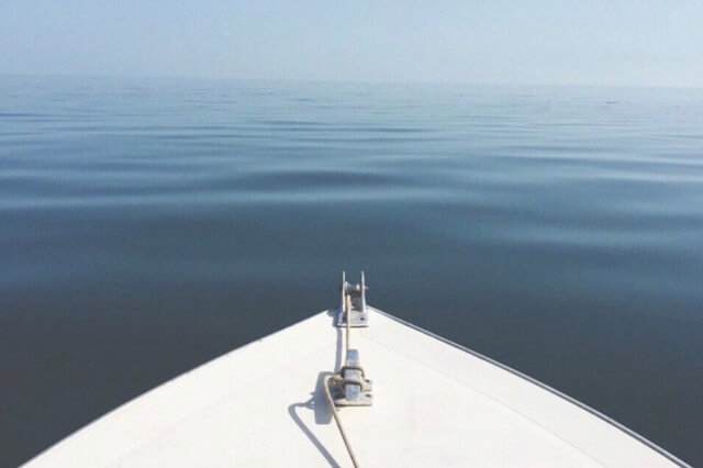View from the bow of a boat heading into a vast, calm ocean under a cloudless, pale blue sky. The water is smooth with gentle ripples, fading into the horizon in perfect stillness.
