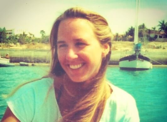 Young woman smiling brightly while sitting on a boat in turquoise water, with a dock, sailboat, and palm trees visible in the sunlit background. Her long blonde hair is gently windswept, and she wears a white shirt under the warm, tropical light.
