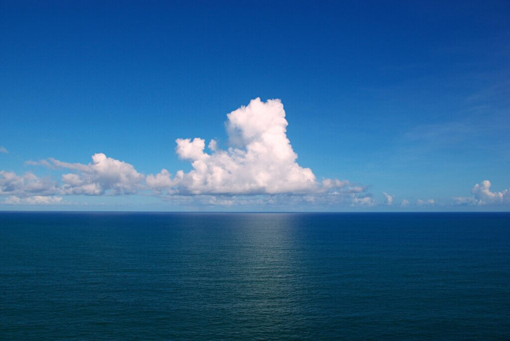 Bright blue sky with a towering cumulus cloud casting a faint reflection over a calm, deep blue ocean. The horizon line is clear, and the cloud's shadow creates a vertical path of lighter water leading straight toward it.