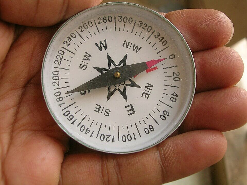 A close-up of a person’s hand holding a compass with a white face, black directional markings, and a red needle pointing north. The background is blurred, keeping focus on the compass and the person’s fingers.