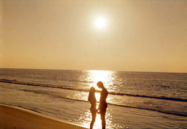 Silhouetted couple standing close together at the edge of the ocean during sunset, with golden sunlight reflecting off the calm waves and casting a warm glow over the beach.