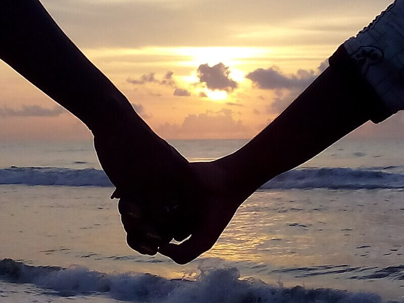 Two people holding hands in silhouette against a golden ocean sunset, with waves gently rolling in and clouds scattered across the glowing sky. The scene captures a peaceful, romantic moment at the beach.