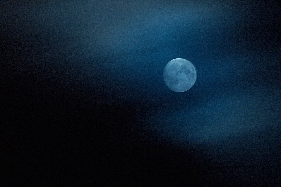 Full moon glowing in a dark, cloudy night sky with soft blue tones. Wisps of thin clouds drift across the scene, partially veiling the moon in a dreamy, ethereal light.