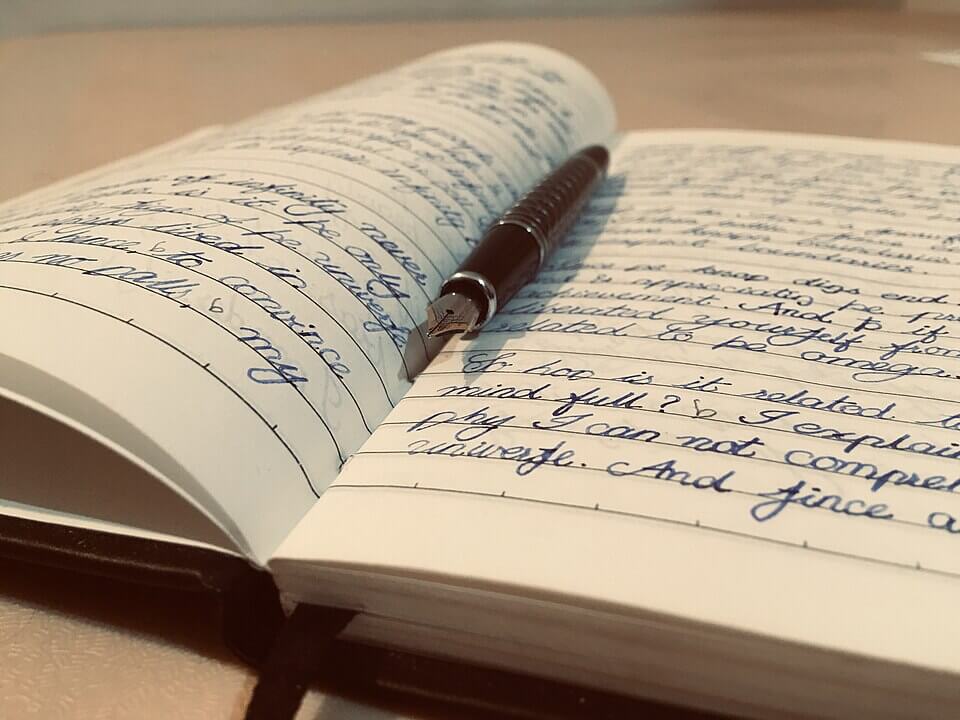 Open notebook filled with neat, cursive handwriting in blue ink, with a fountain pen resting across the pages. The scene is warmly lit, and the focus highlights the pen's metallic nib and the texture of the paper.