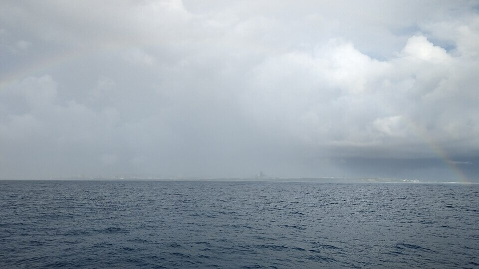 Vast expanse of ocean under a cloudy sky, with a faint rainbow arching on the right side of the image and a barely visible coastline in the distance. The water appears calm with small ripples, and the sky is overcast with shades of gray and white.