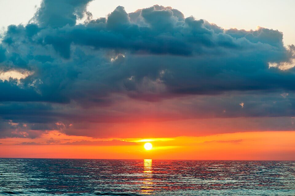 Vivid ocean sunset with the sun glowing just above the horizon, casting golden and orange reflections on the water below. Dramatic, dark clouds hang overhead, contrasting with the warm, fiery tones of the sky.