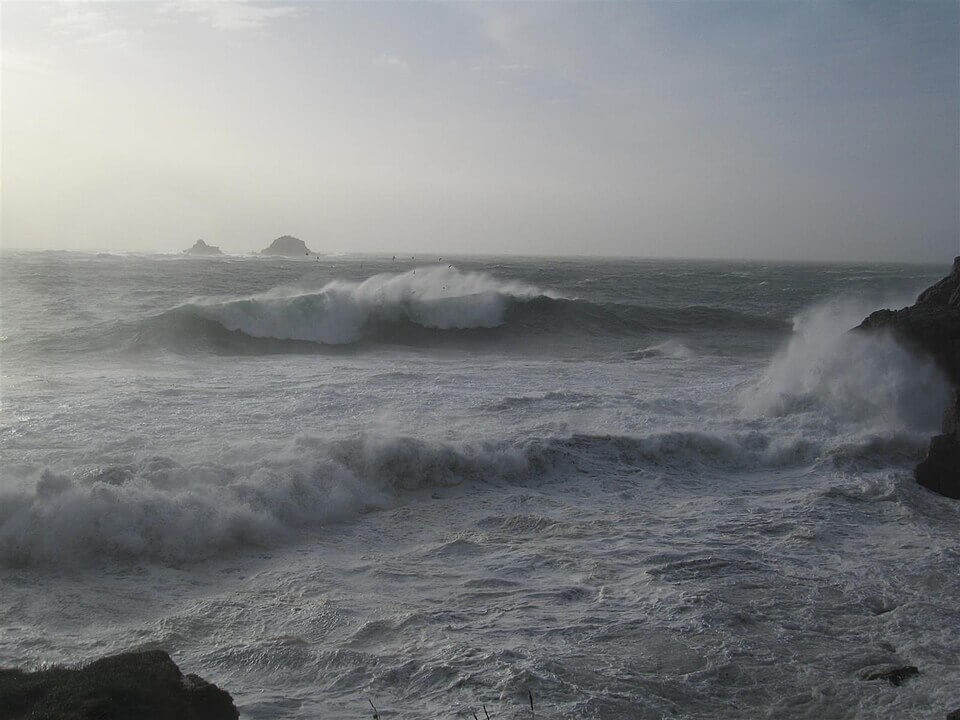 Large, foamy waves crash violently against a rocky shoreline under a hazy, gray sky. The ocean appears turbulent and stormy, with sea spray and distant rock formations visible on the horizon.