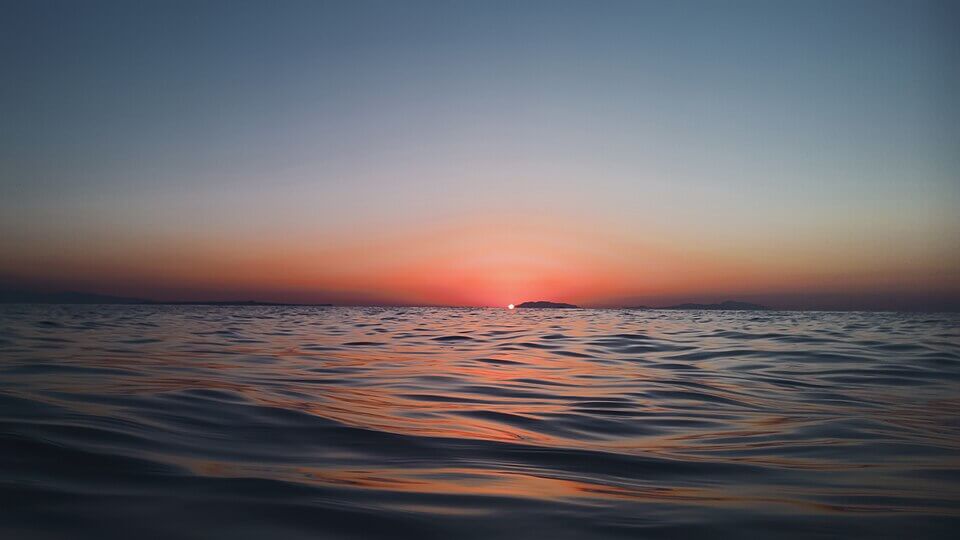 Tranquil ocean at sunset with small ripples on the surface, reflecting warm hues of orange, pink, and purple. The sun is just above the horizon, casting a soft glow across the distant silhouette of low-lying islands.