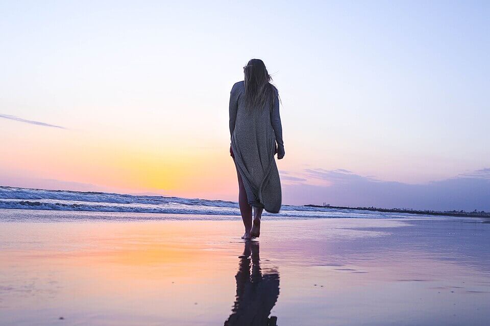 Woman in a long gray dress walks barefoot along a beach at sunset, her reflection faintly mirrored in the wet sand. The sky is painted in soft purples, pinks, and oranges as waves gently roll in.