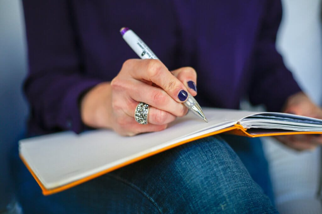 Close-up of a person writing in a lined notebook with a pen, wearing a sparkly statement ring and dark purple nail polish. The person is dressed in a purple top and blue jeans, with the notebook resting on their lap.